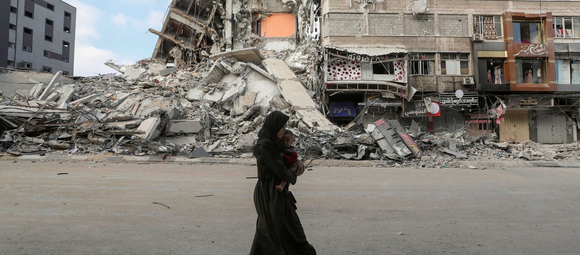 امرأة تحمل طفلها تسير على خلفية ركام منازل، تخلو حييها بعد اشتداد قصف المدافع والطيران الحربي الإسرائيلي في شمال قطاع غزة، فلسطين 14 مايو 2021 - سبوتنيك عربي, 1920, 21.05.2021