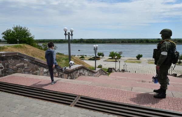 جندي روسي يقوم بدورية بينما صبي يلعب كرة القدم على ضفة نهر في خيرسون، وسط العملية العسكرية الروسية المستمرة في أوكرانيا،  20 مايو 2022 - سبوتنيك عربي