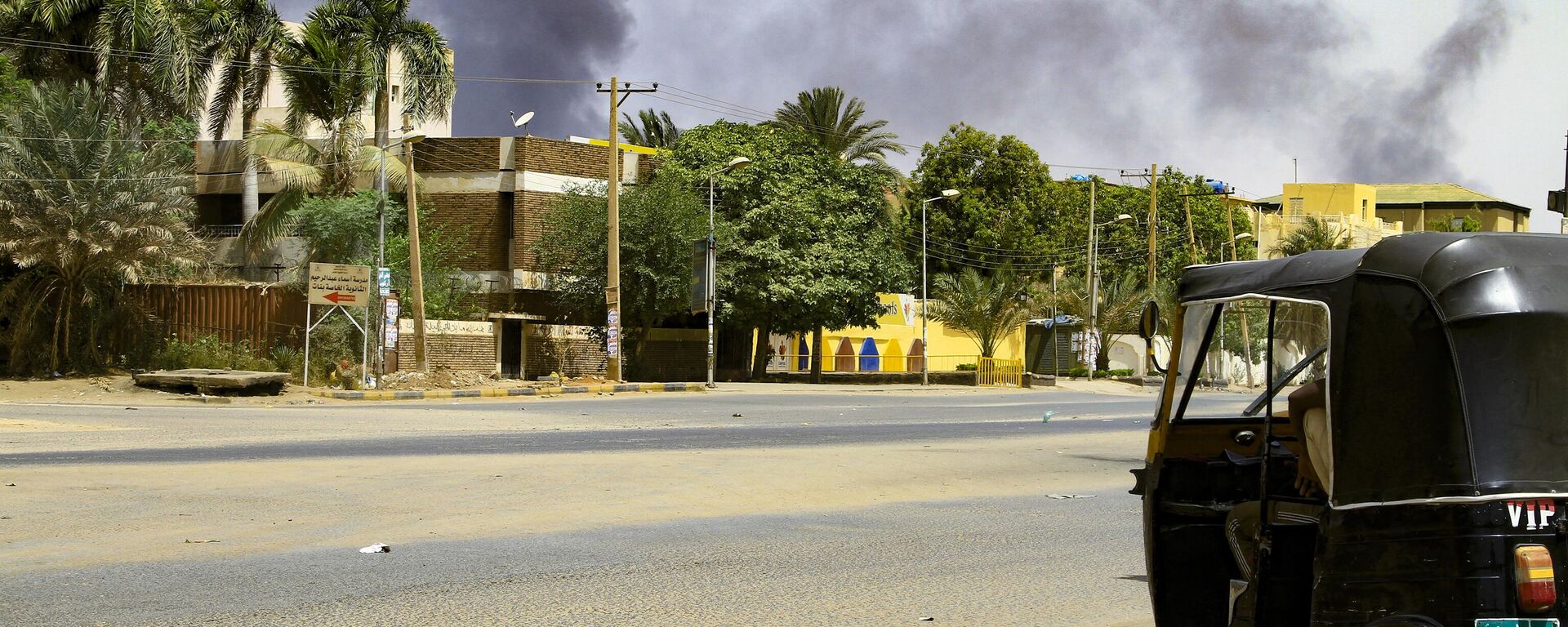 الدخان يتصاعد من المباني بينما يجلس سائق تاكسي التوكتوك في سيارته على طول شارع مهجور في الخرطوم في 16 أبريل 2023 ، أثناء القتال المستمر بين قوات اثنين من الجنرالات المتناحرين. - سبوتنيك عربي, 1920, 19.04.2023
