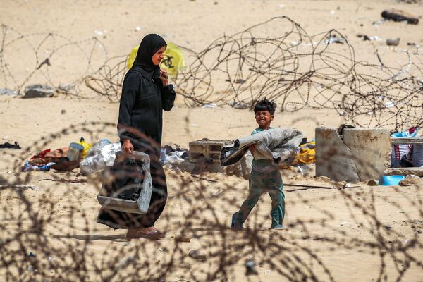 امرأة وصبي يسيران مع أمتعتهما عبر سياج الأسلاك الشائكة أثناء نزوحهما من رفح جنوبي قطاع غزة في 11 مايو 2024 - سبوتنيك عربي