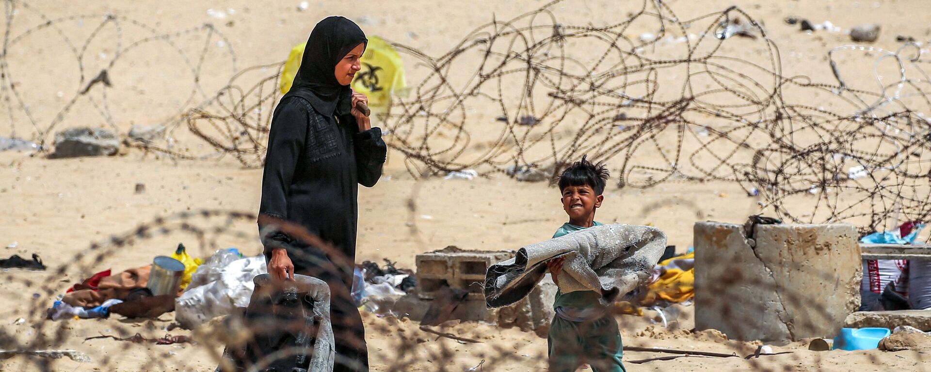 امرأة وصبي يسيران مع أمتعتهما عبر سياج الأسلاك الشائكة أثناء نزوحهما من رفح جنوبي قطاع غزة في 11 مايو 2024 - سبوتنيك عربي, 1920, 18.05.2024