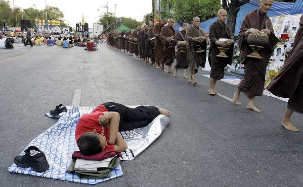 رهبان بوذيون يصطفون للحصول على الصدقات الصباحية أمام متظاهر نائم في الشارع بعد ليلة من المظاهرات خارج مقر الحكومة في بانكوك في تايلاند - سبوتنيك عربي