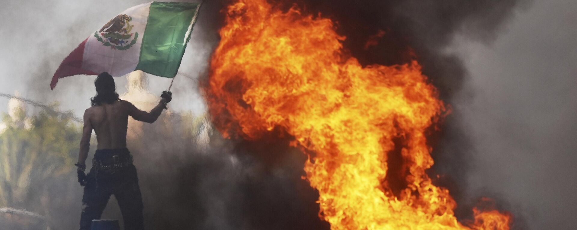 Протестующий машет мексиканским флагом на фоне горящего автомобиля - سبوتنيك عربي, 1920, 09.06.2025