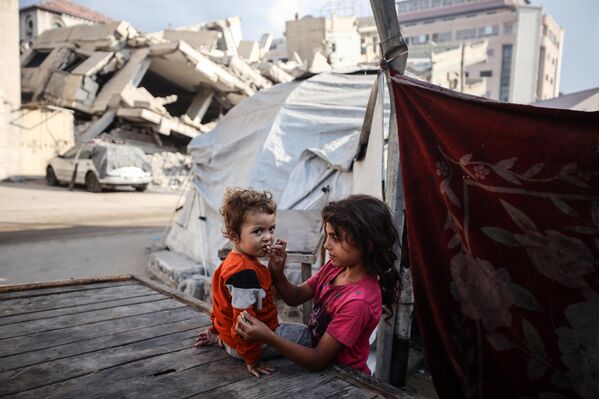 فتاة فلسطينية نازحة تطعم أختها الخبز أمام مدرسة الفارابي، التي تحولت إلى ملجأ بعد تعرضها لغارة إسرائيلية، في مدينة غزة. - سبوتنيك عربي