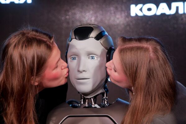 صورة تظهر الروبوت &quot;أيدول&quot; ينظر للأمام، بينما يقبله شخصان من جانبي وجهه، ما يعكس التفاعل البشري مع هذا الروبوت. - سبوتنيك عربي