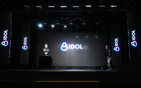 مشهد من عرض تقديمي في قاعة مظلمة، حيث يقف الروبوت &quot;أيدول&quot; على منصة أمام شاشة كبيرة تحمل شعاره، ويحيط به شعارات &quot;أيدول&quot; على جانبي المسرح. - سبوتنيك عربي