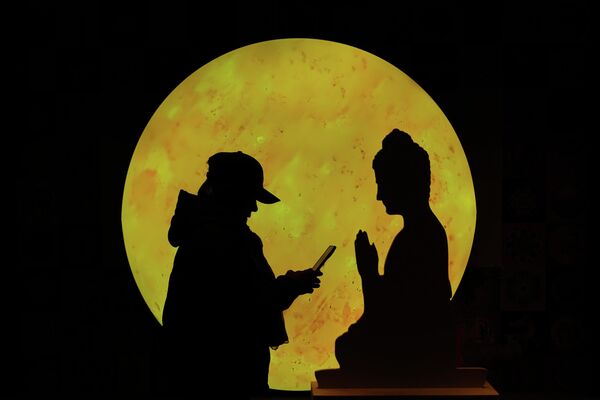 امرأة تتصفح هاتفها الذكي أمام قطعة من الورق المقوى على شكل بوذا مقابل قمر معروض في مركز للتسوق، في بكين. - سبوتنيك عربي