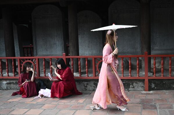 صورة تظهر فتيات، في ساحة معبد الأدب في هانوي ، في فيتنام، وتظهر فتاتان مرتديتان رداء أحمر اللون، وفتاة أخرى في فستان زهري اللون تسير حاملة مظلة. - سبوتنيك عربي