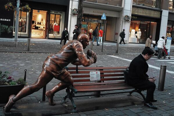 صورة تظهر رجلا في لباس أسود اللون، يجلس على مقعد خشبي، وبجوار تمثال  رجل رياضي ويمسك بيده كرة طائرة، الصورة تم التقاطها في شارع تجاري في حي غينزا، في طوكيو. - سبوتنيك عربي