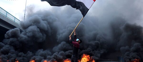 متظاهر يحمل العلم العراقي بجانب الإطارات المحترقة أثناء الاحتجاجات المستمرة المناهضة للحكومة في كربلاء - سبوتنيك عربي