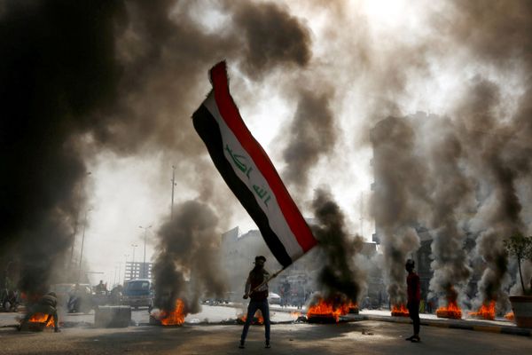 متظاهر يمسك بالعلم العراقي وسط دخان يتصاعد من الإطارات المشتعلة أثناء الاحتجاجات في النجف، العراق 26 نوفمبر 2019 - سبوتنيك عربي