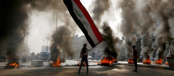 متظاهر يمسك بالعلم العراقي وسط دخان يتصاعد من الإطارات المشتعلة أثناء الاحتجاجات في النجف، العراق 26 نوفمبر 2019 - سبوتنيك عربي