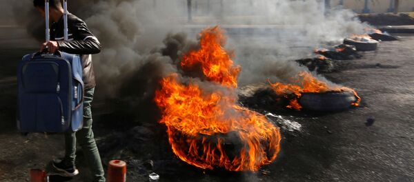 راكب يحمل حقيبته وهو يمشي عبر إطارات محترقة أشعلها المحتجون العراقيون لإغلاق الطريق المؤدي إلى مطار النجف الدولي خلال الاحتجاجات المستمرة المناهضة للحكومة - سبوتنيك عربي