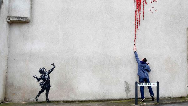 أحد المارة يقف على خلفية رسم غرافيتي لفنان الشارع الشهير بانكي، الرسم لفتاة صغيرة تطلق مقلاعًا من الزهور، على جدار مبنى في بريستول، بريطانيا 14 فبراير 2020. - سبوتنيك عربي